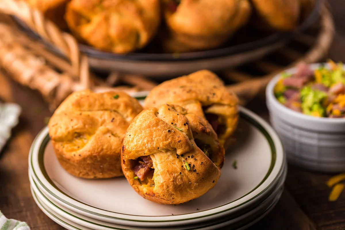 Three biscuit muffin cups with visible meat and cheese filling are served on stacked plates, with a blurred tray and bowl of toppings in the background.
