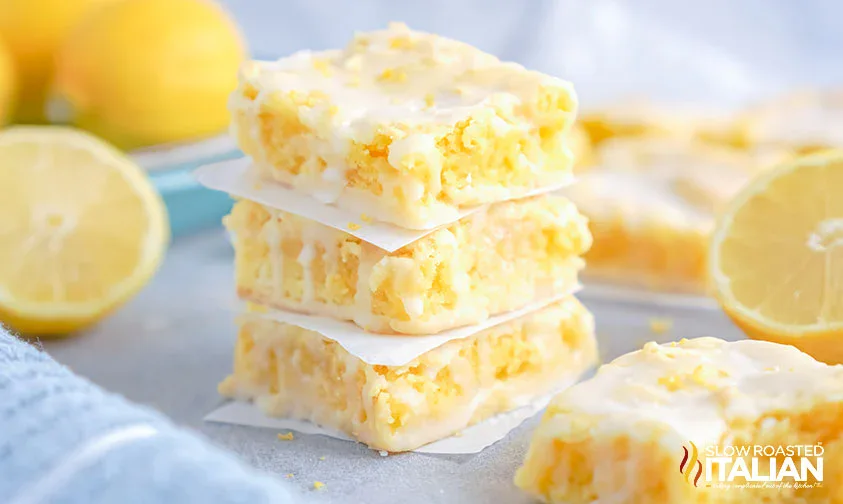 A stack of three glazed lemon bars separated by parchment paper, with lemons and more lemon bars in the background.