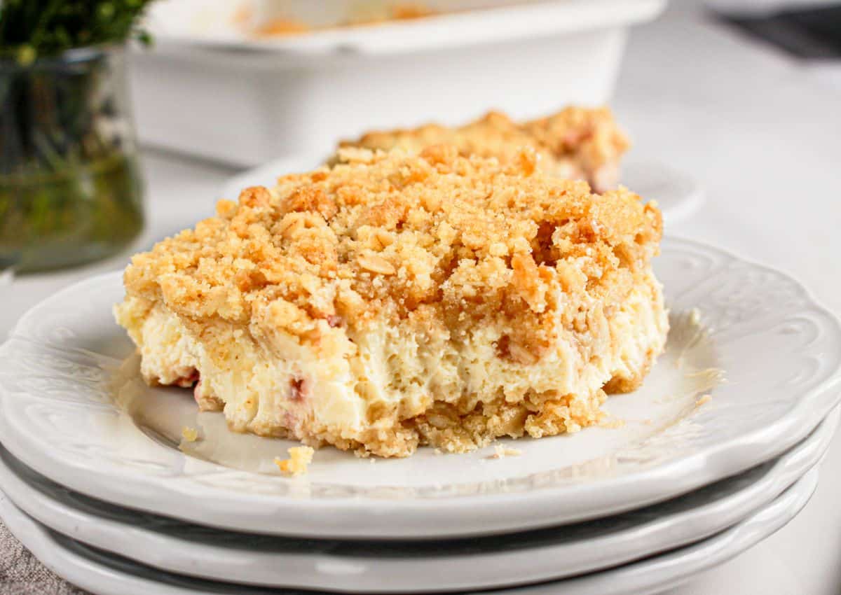 A slice of creamy dessert with a crumbly topping sits on a white plate, with another plate and a baking dish in the background.