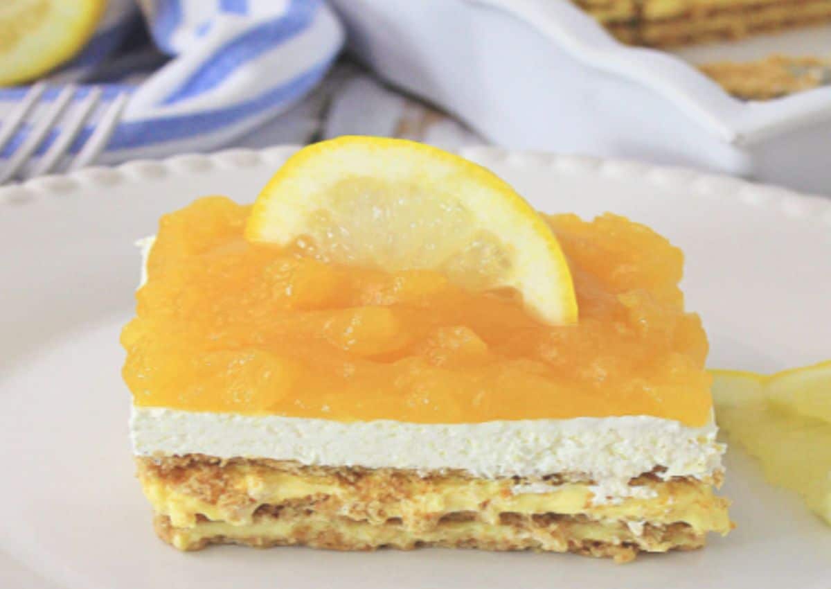 A layered dessert bar with a graham cracker base, creamy filling, orange topping, and a lemon slice garnish on a white plate.
