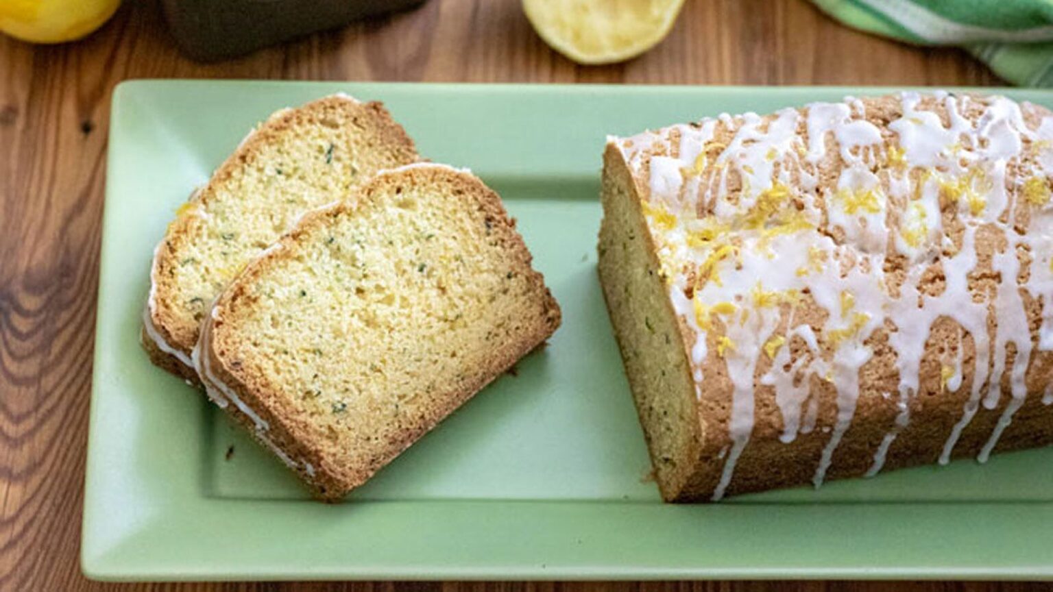 A loaf of glazed bread with two slices cut, displayed on a green rectangular plate on a wooden surface.