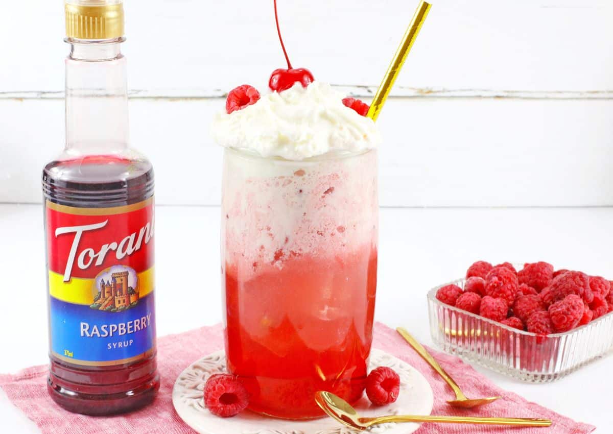 A glass of raspberry Italian soda topped with whipped cream and a cherry, next to a bottle of Torani raspberry syrup and a bowl of fresh raspberries on a pink cloth.