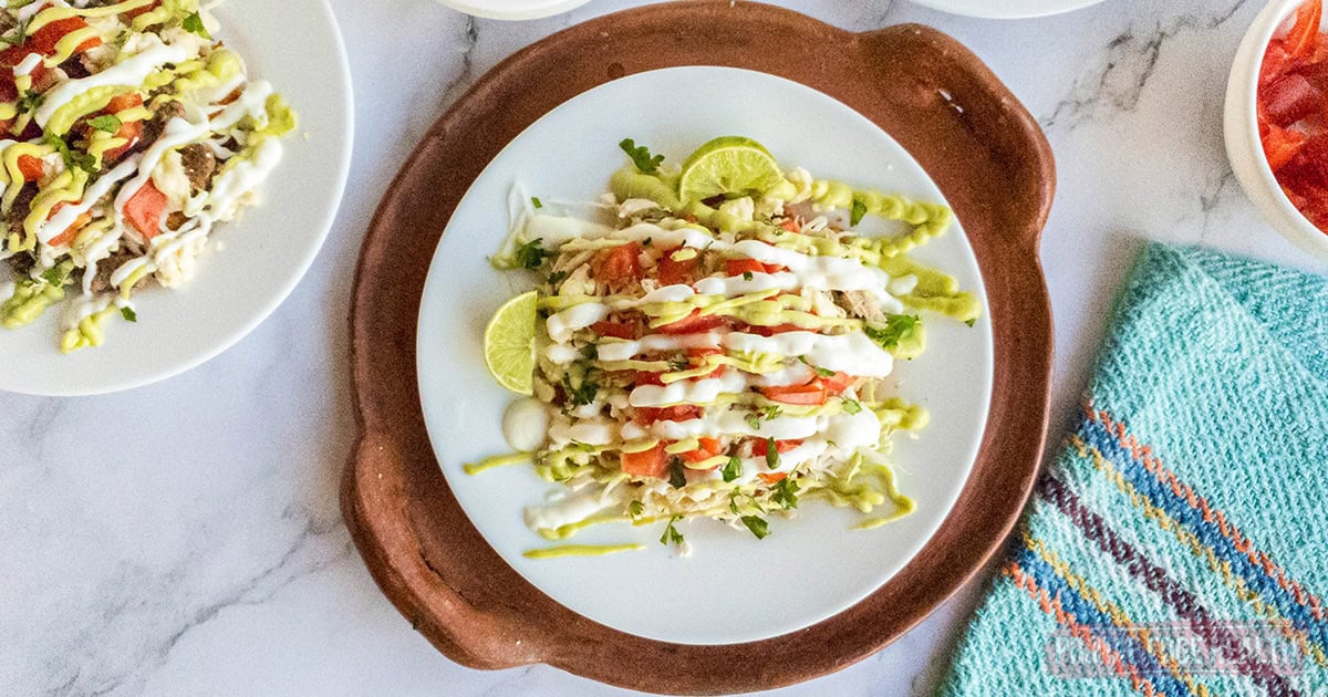 A plate of tostadas topped with shredded lettuce, diced tomatoes, lime wedges, and drizzled with white and green sauces, seen from above on a white table with a blue-striped cloth nearby.