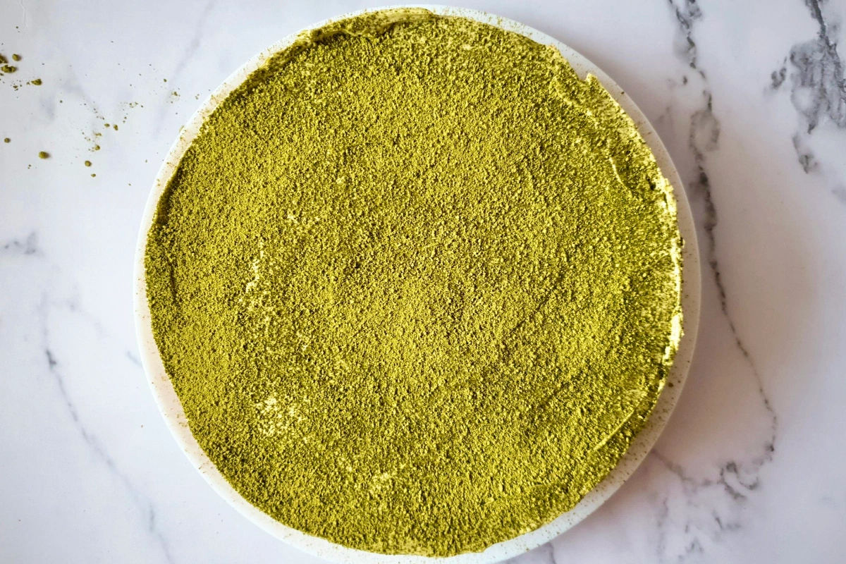 A round cake with a smooth, even layer of green matcha powder on top, placed on a white marble surface.