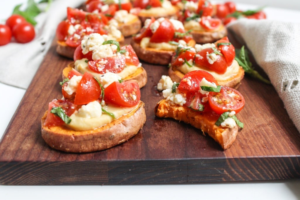 Slices of roasted sweet potato topped with hummus, cherry tomatoes, crumbled cheese, and herbs on a wooden board; one slice has a bite taken out.