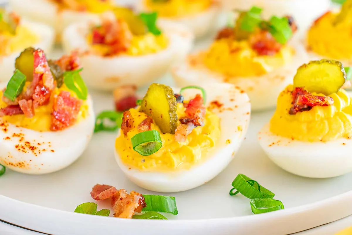 Deviled eggs topped with sliced pickles, chopped bacon, and green onions are arranged on a white plate.