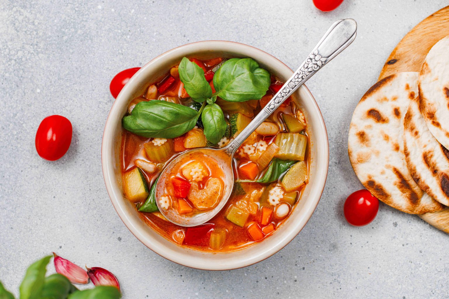 A bowl of vegetable soup with a spoon, garnished with fresh basil, surrounded by cherry tomatoes and flatbread on a light surface.