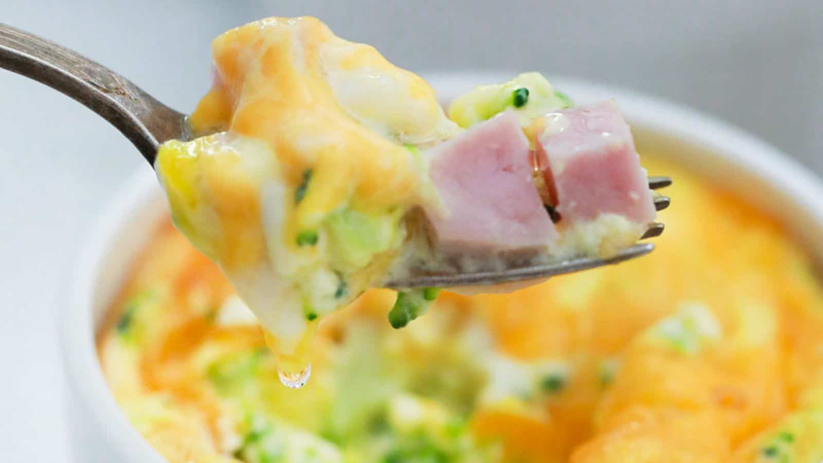 A fork holds a bite of baked casserole with chunks of ham, melted cheese, broccoli, and egg, with more of the casserole visible in a dish below.