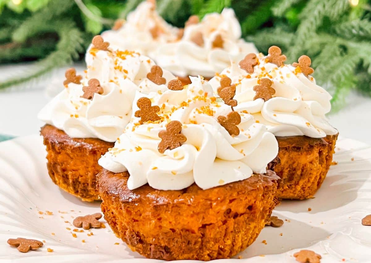 Mini cheesecakes topped with swirls of whipped cream and small gingerbread man sprinkles are arranged on a white plate with a green leafy background.