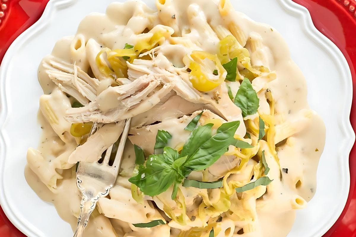 Pasta with shredded chicken, creamy white sauce, sliced pepperoncini, and fresh basil garnish on a white plate with a fork.
