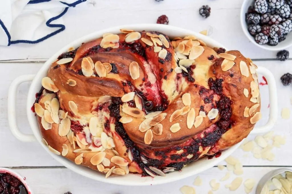 Oval dish with braided bread filled with berry jam and topped with sliced almonds, placed on a white wooden surface with scattered blackberries and almond slices nearby.
