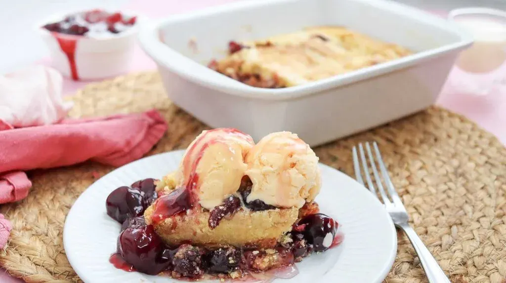 A slice of cherry cobbler topped with two scoops of vanilla ice cream on a white plate, with a fork and a baking dish of cobbler in the background.