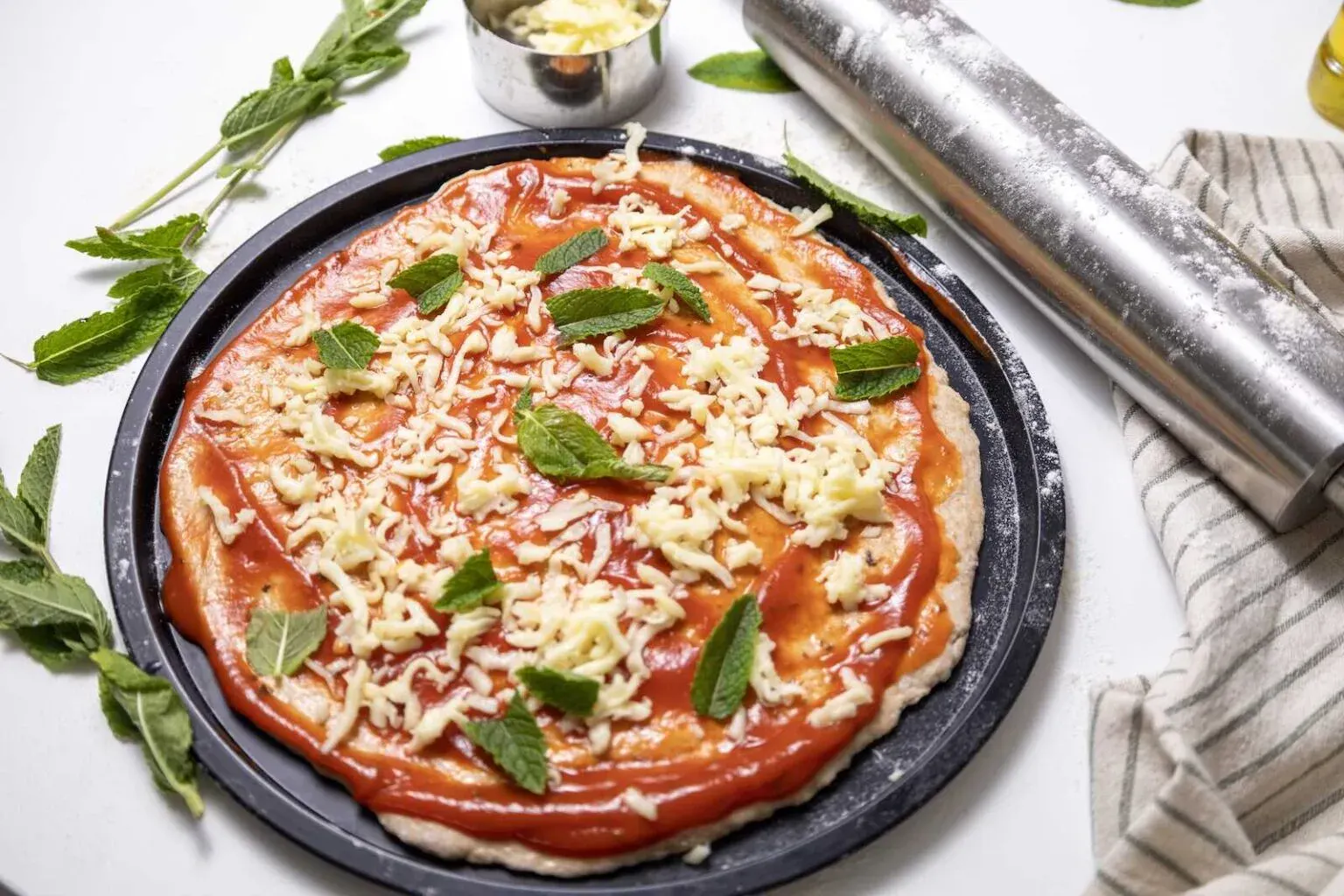 Unbaked pizza on a round tray topped with tomato sauce, shredded cheese, and mint leaves. A rolling pin, a cup of cheese, and fresh mint are nearby on a white surface.