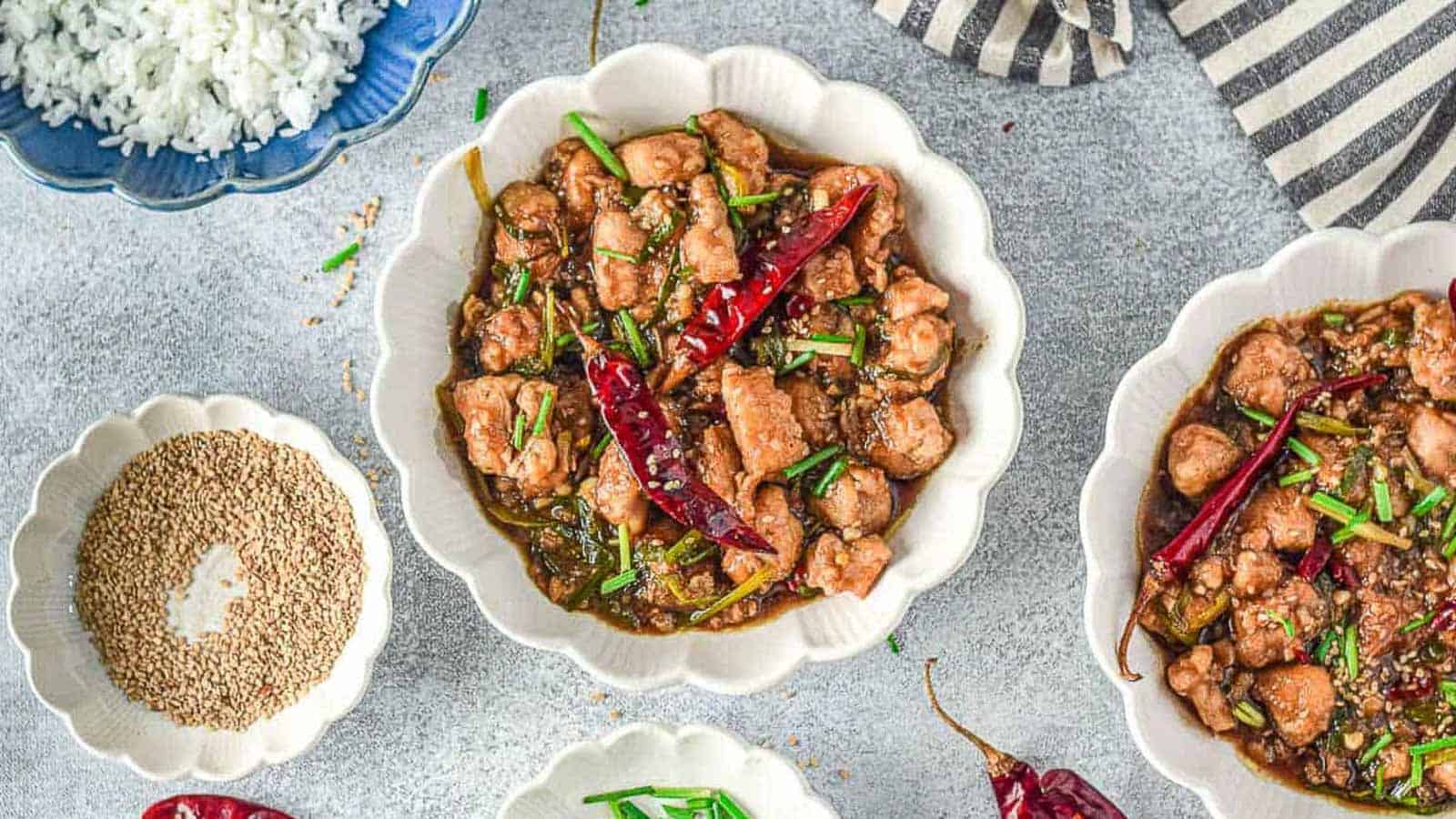 Bowls of cooked chicken with dried red chilies and green herbs, served alongside a bowl of white rice and a bowl of sesame seeds on a gray surface.