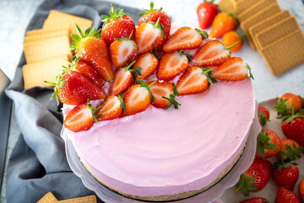 A pink strawberry cheesecake topped with halved fresh strawberries sits on a cake stand, surrounded by whole strawberries and rectangular biscuits.