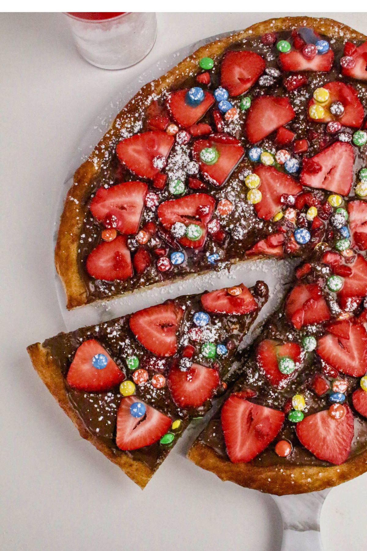 A dessert pizza topped with sliced strawberries, colorful chocolate candies, and powdered sugar, with one slice being removed.