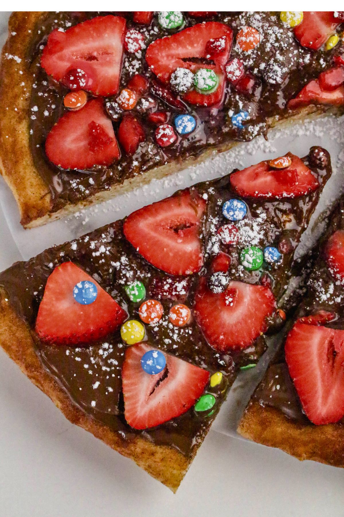 A dessert pizza topped with chocolate spread, fresh strawberry slices, colorful candy-coated chocolates, and a dusting of powdered sugar.