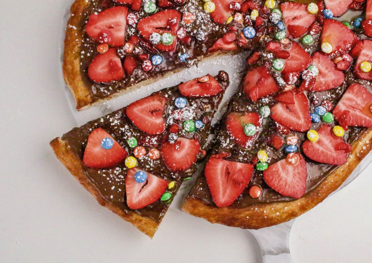 A dessert pizza topped with chocolate spread, sliced strawberries, colorful candy-coated chocolates, and powdered sugar, with one slice being removed.