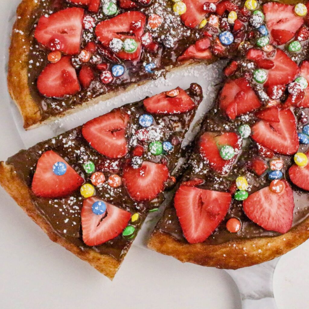 A dessert pizza topped with chocolate spread, sliced strawberries, colorful candy-coated chocolates, and powdered sugar, with one slice being lifted from the whole.