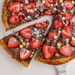 A dessert pizza topped with chocolate spread, sliced strawberries, colorful candy-coated chocolates, and powdered sugar, with one slice being lifted from the whole.