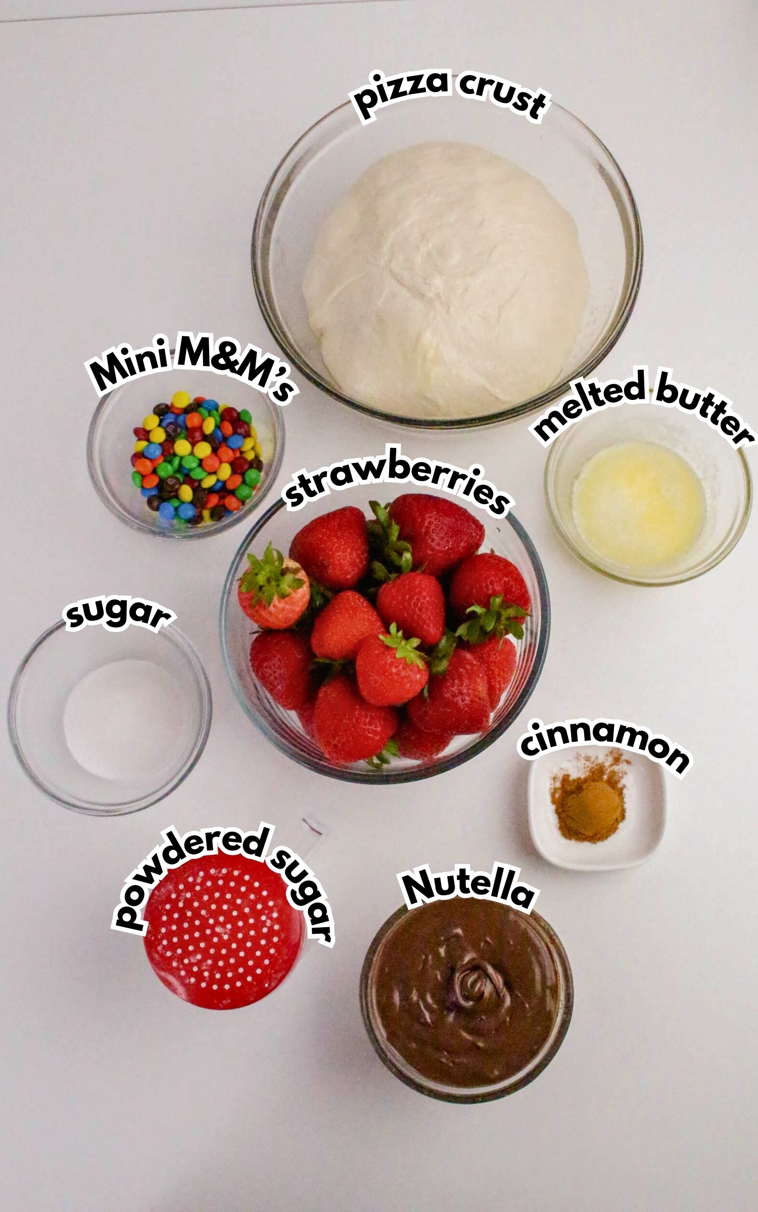 Pizza crust dough, melted butter, strawberries, Nutella, powdered sugar, sugar, cinnamon, and mini M&M’s arranged in separate clear bowls on a white surface.