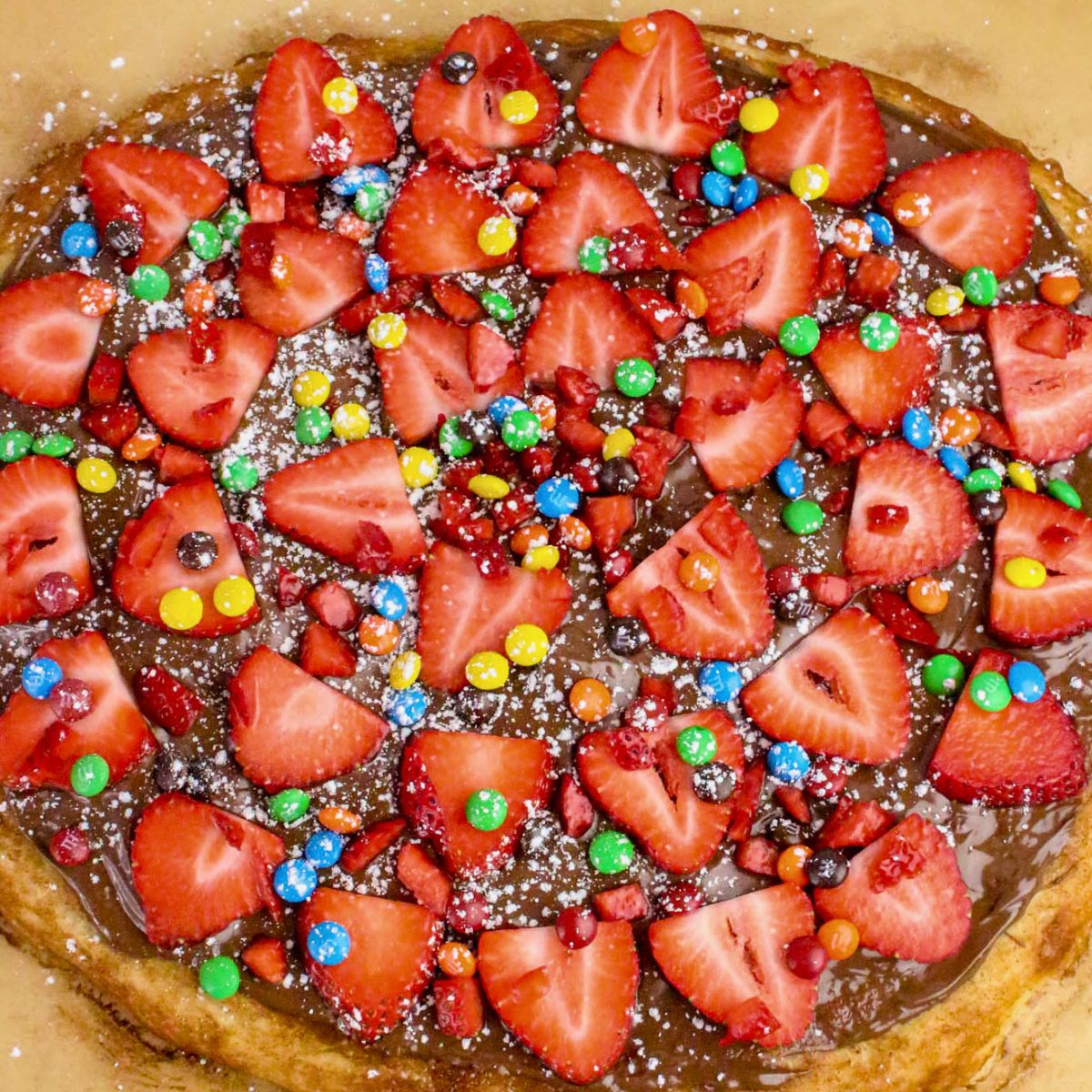 A delicious Nutella dessert pizza topped with fresh strawberry slices, colorful candy pieces, and a dusting of powdered sugar.