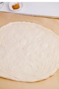 A rolled-out round pizza dough on parchment paper with a small dish of cinnamon on the counter in the background.