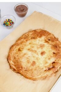 A large baked pastry on parchment paper is shown with small bowls of chocolate spread and colorful candy nearby on a white surface.