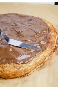 An offset spatula spreads melted chocolate over a round baked pastry on parchment paper.