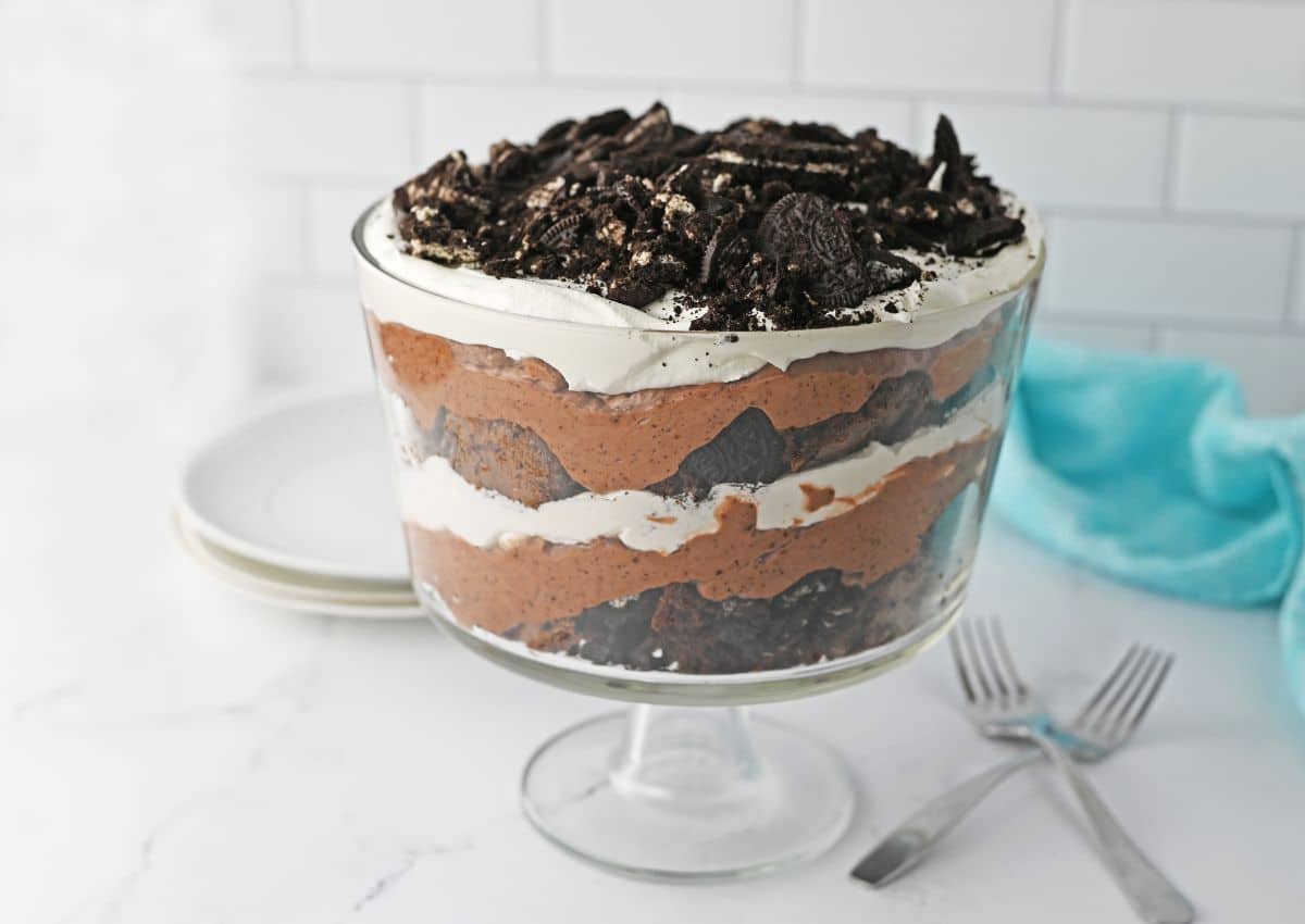 A glass trifle dish filled with layered chocolate mousse, whipped cream, chocolate cake, and crushed cookies, with plates and forks beside it on a white surface.