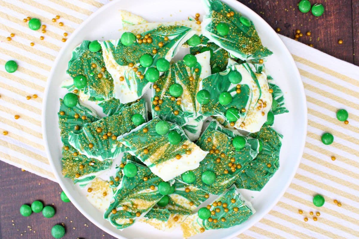 A white plate with pieces of green and white chocolate bark topped with green candy balls and gold sprinkles, placed on a striped cloth.