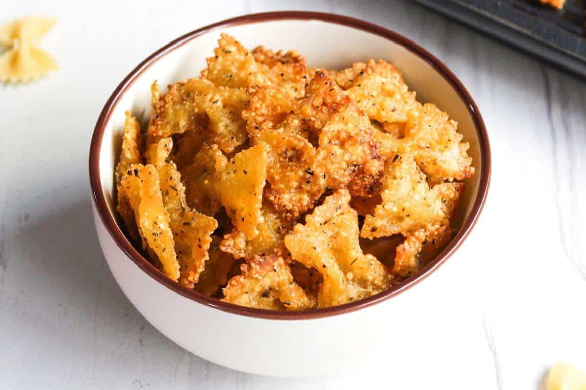 A white bowl filled with crispy, seasoned bow tie pasta chips on a white surface.