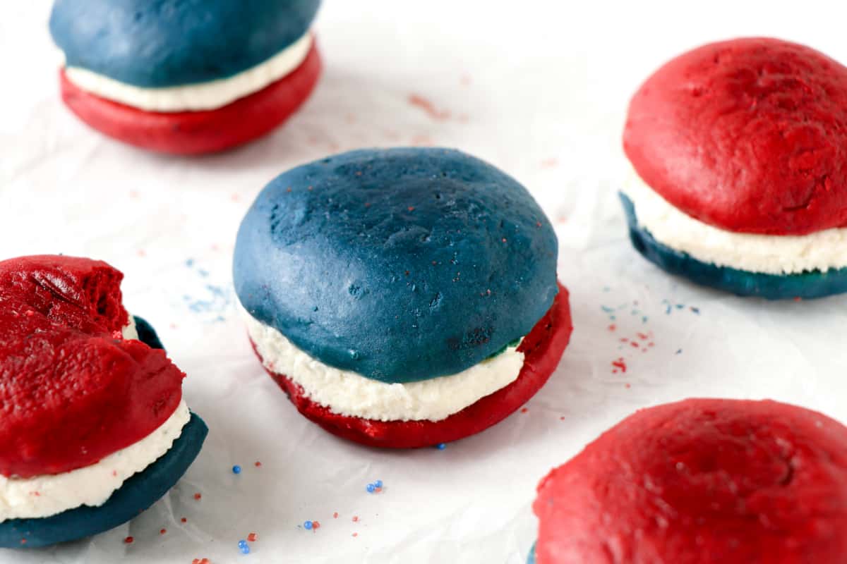 Red and blue whoopie pies with white cream filling are arranged on a white surface; one has a bite taken out of it.