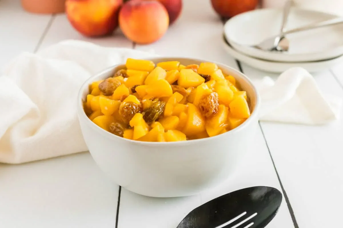 A white bowl filled with diced peaches and golden raisins sits on a white table next to a black serving spoon, with whole peaches and plates in the background.