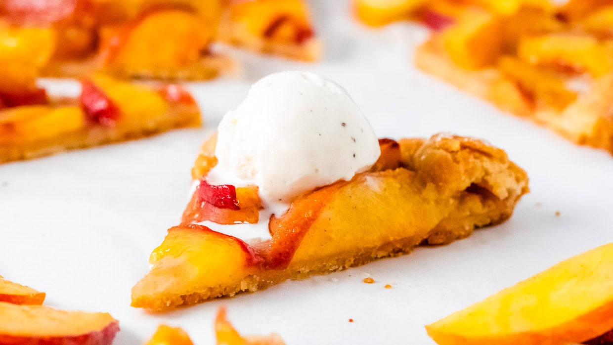 A slice of peach tart with a scoop of vanilla ice cream on top, sitting on a white surface with peach slices scattered around.