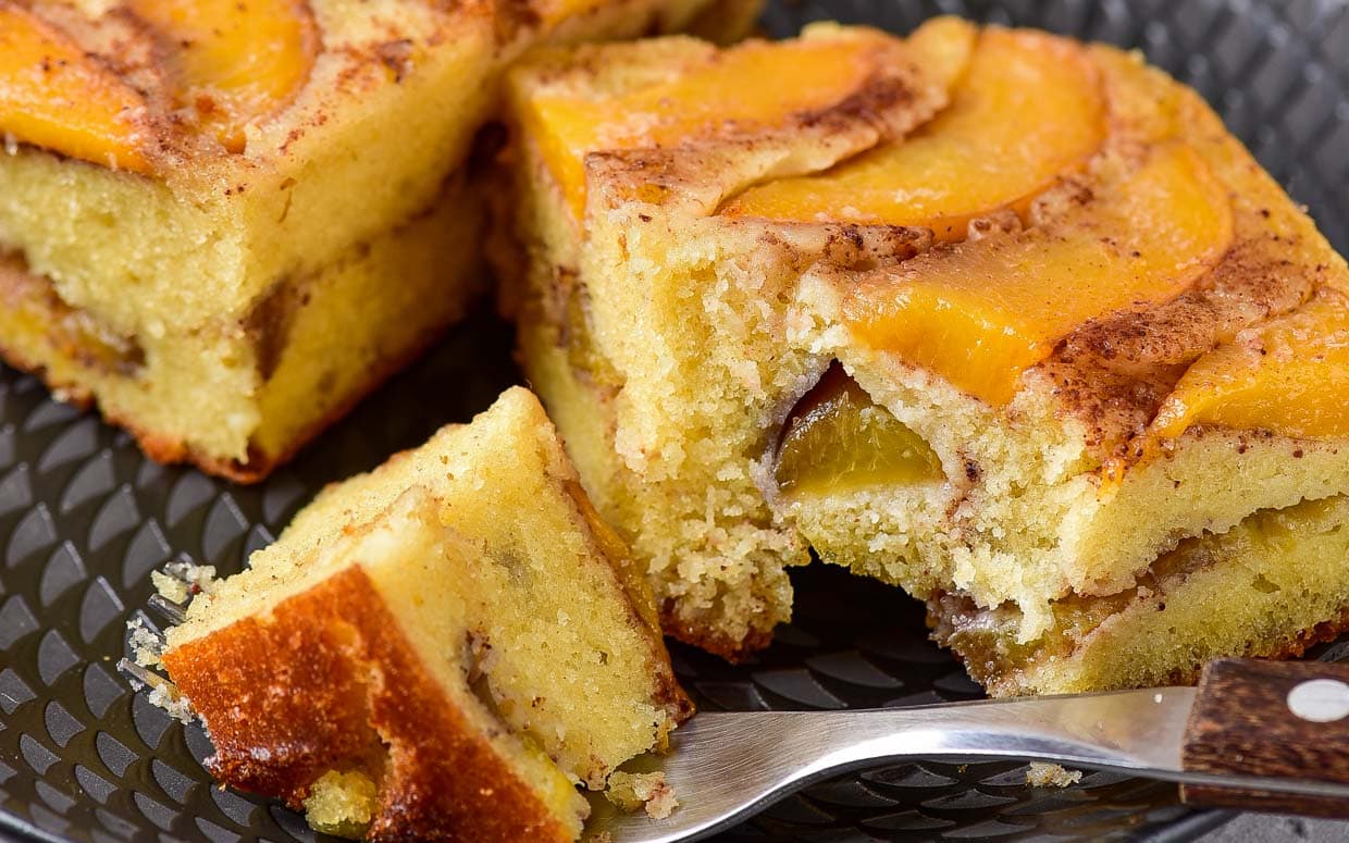 Close-up of two slices of peach cake on a dark plate, with a fork holding a small piece; the cake shows layers of peaches and a moist, yellow interior.