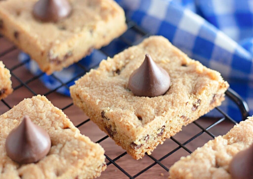 A cooling rack with rectangular cookie bars, each topped with a single chocolate kiss, and a blue and white checkered cloth in the background.