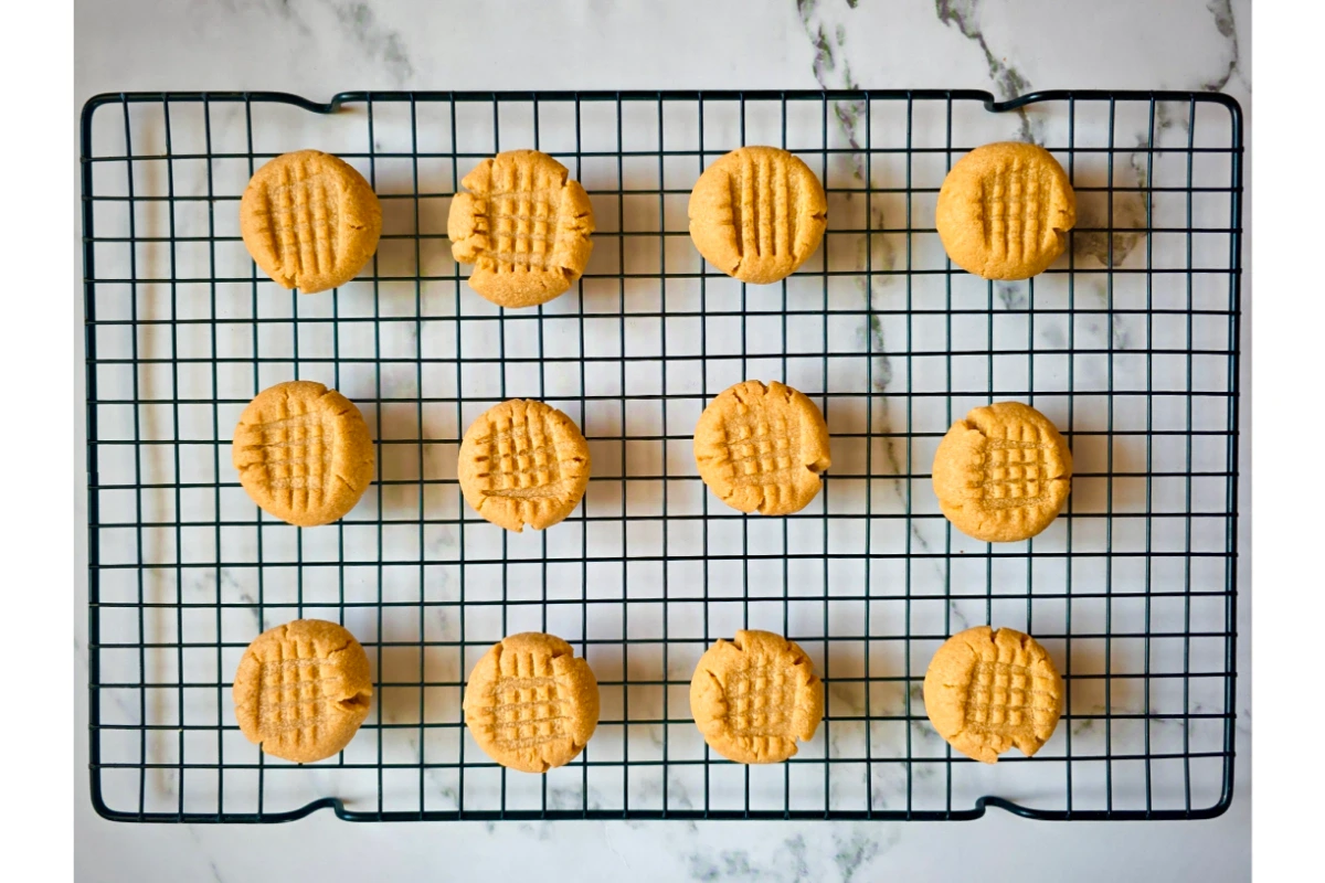 A cooling rack with fourteen round peanut butter cookies arranged in rows, each with a crisscross fork pattern on top, on a marble surface.