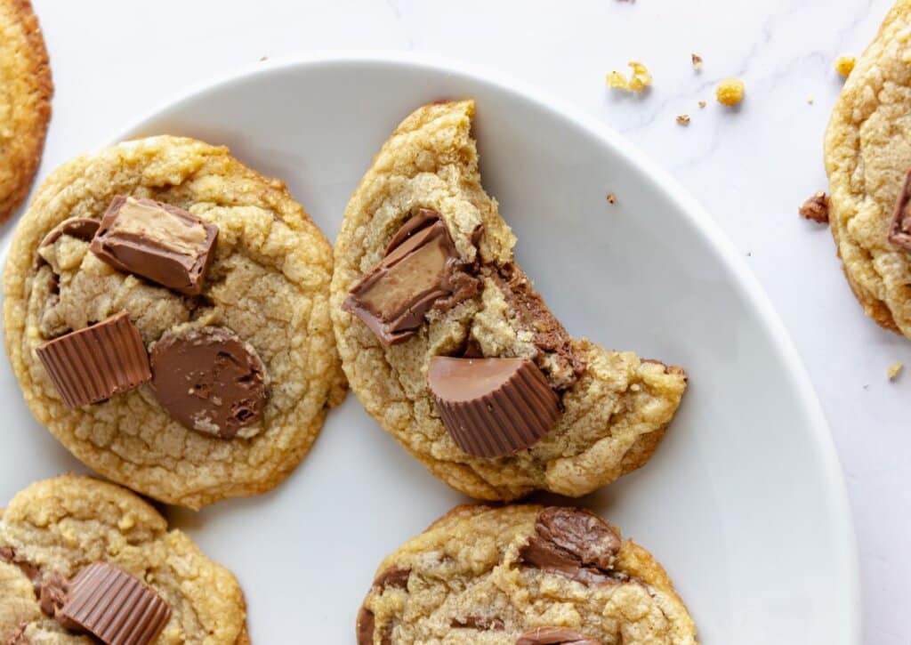 A plate of peanut butter cup cookies; one cookie has a bite taken out, and others are topped with chocolate peanut butter cup pieces.