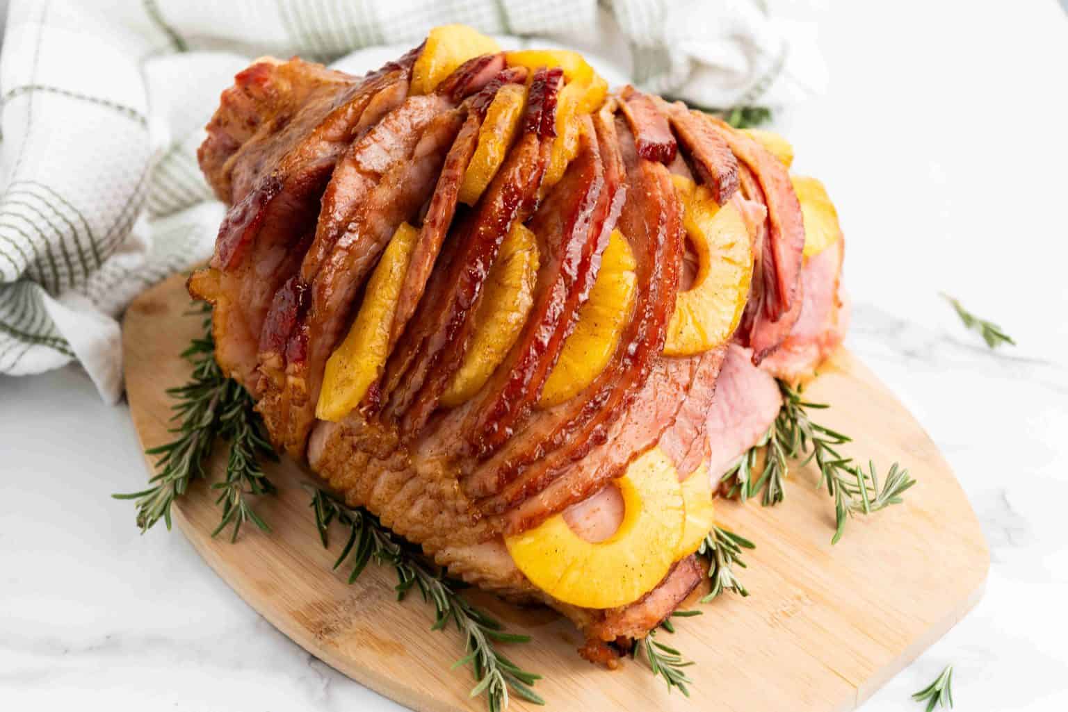 Sliced baked ham garnished with pineapple rings and cherries, served on a wooden board with sprigs of rosemary.