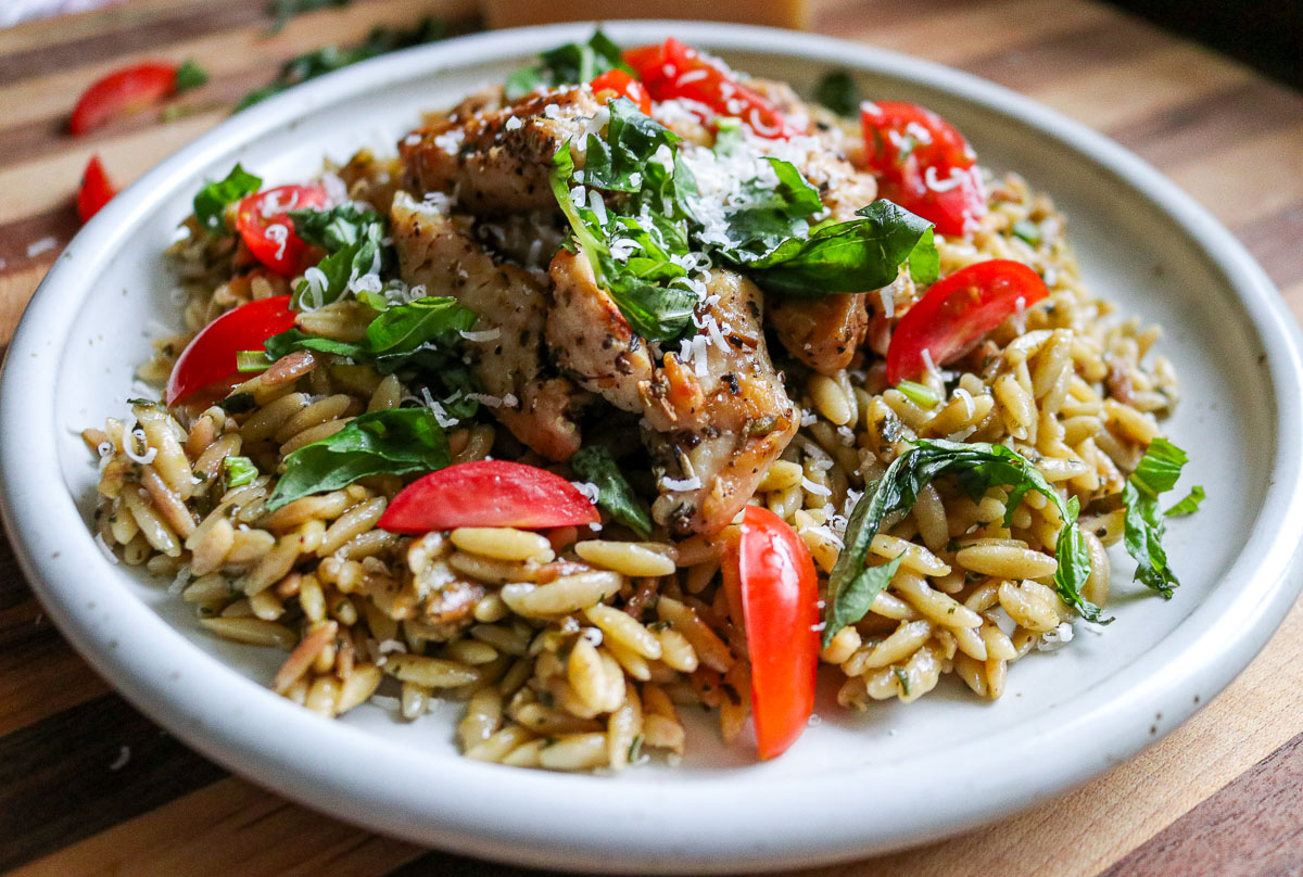 A plate of orzo pasta topped with grilled chicken, cherry tomato slices, fresh basil, and grated cheese on a wooden surface.