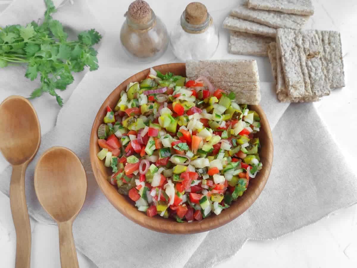 A wooden bowl of chopped vegetable salad with crispbread, wooden spoons, cilantro, salt and pepper shakers on a cloth napkin.