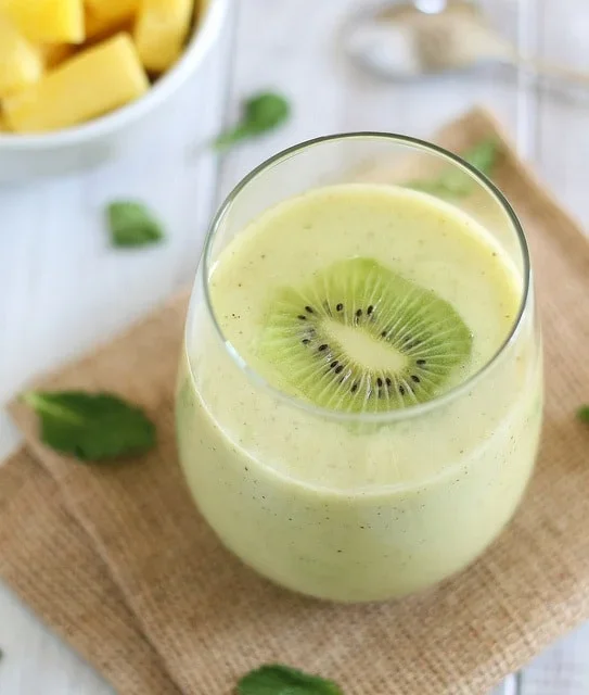 A glass of green smoothie with a slice of kiwi on top, placed on a burlap mat, with mint leaves and a bowl of pineapple pieces in the background.