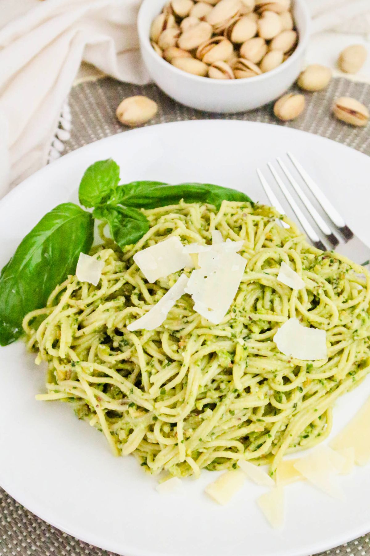 Plate of spaghetti with green pesto sauce, garnished with basil leaves and shaved parmesan, with a bowl of pistachios in the background.