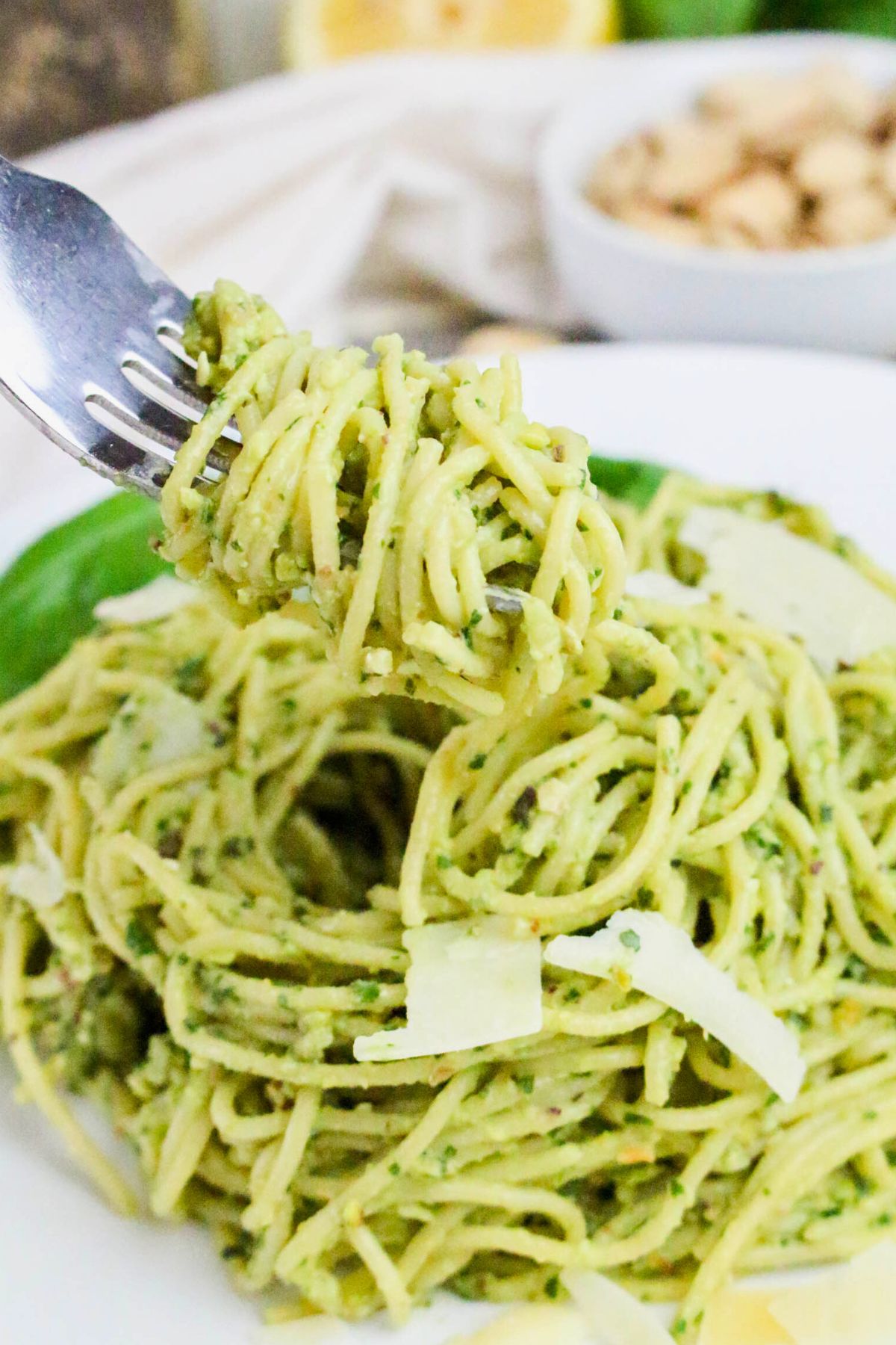 A fork lifts a serving of spaghetti coated in green pesto sauce, with parmesan cheese shavings on top, on a white plate.
