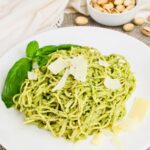 A plate of spaghetti with green pesto sauce, garnished with basil leaves and parmesan cheese, with a bowl of pistachios in the background.