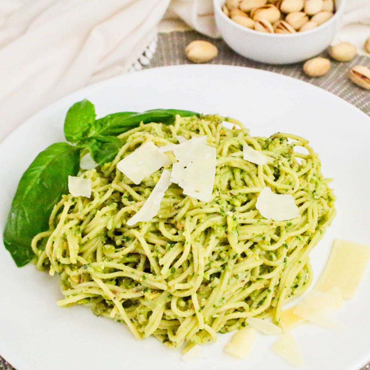 A plate of spaghetti with green pesto sauce, garnished with basil leaves and parmesan cheese, with a bowl of pistachios in the background.