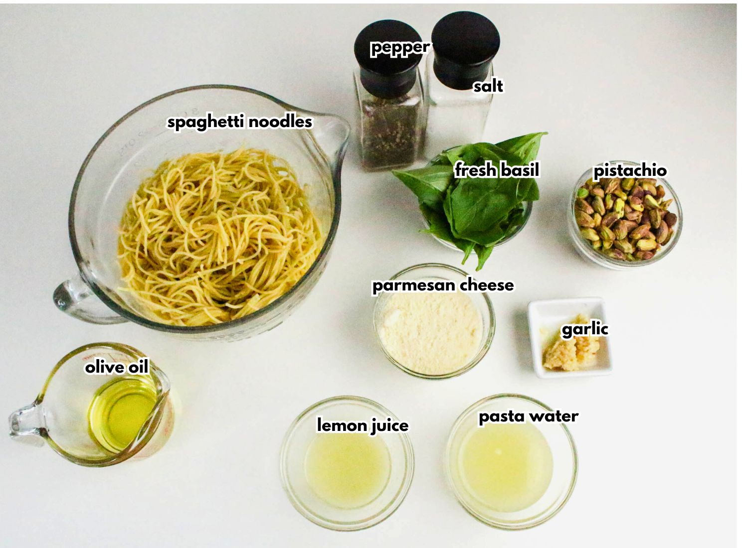Ingredients for pesto pasta are arranged on a white surface, including spaghetti noodles, olive oil, lemon juice, pasta water, garlic, parmesan cheese, pistachios, fresh basil, salt, and pepper.