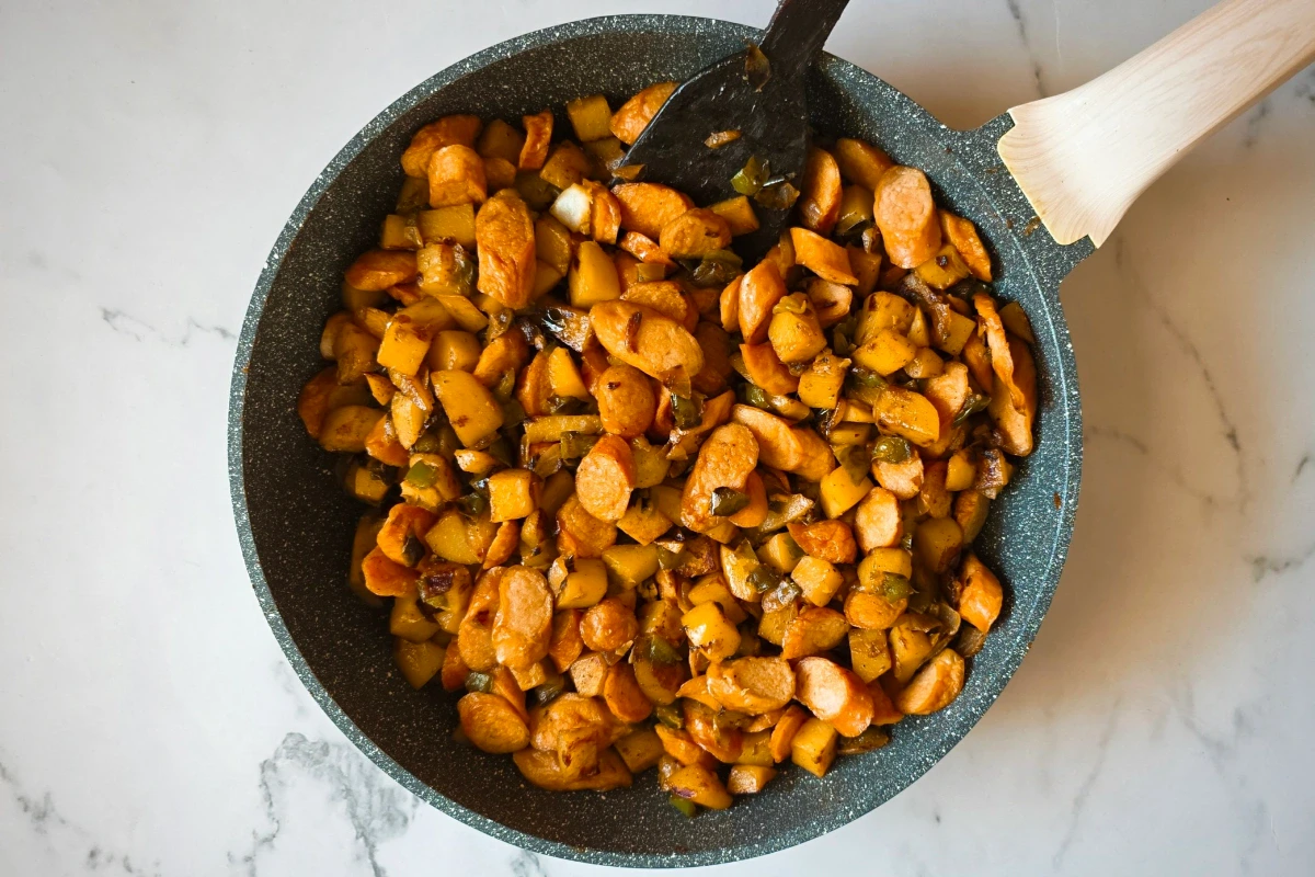 A pan filled with sautรฉed sliced carrots and chopped vegetables, stirred with a black spatula, sits on a light marble surface.