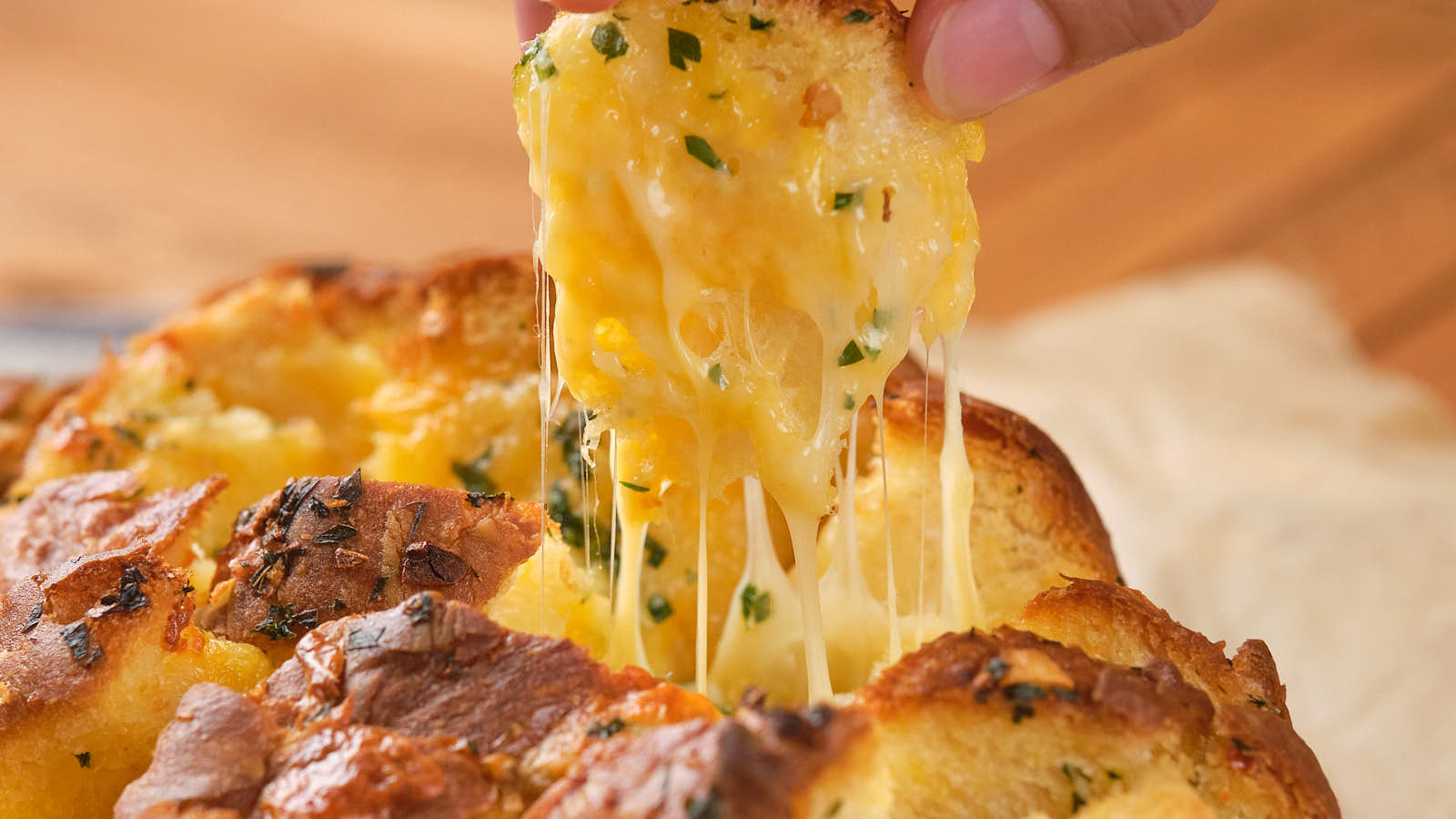 A hand pulls a piece of cheesy garlic bread, showing melted cheese stretching from the loaf.